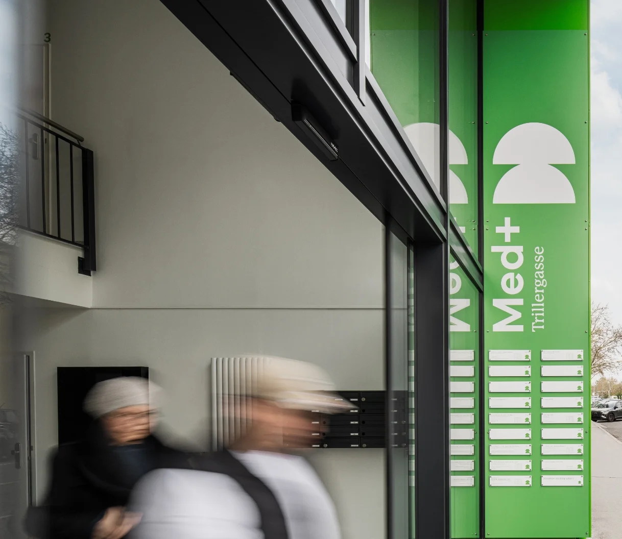 Eingang zum Med+ Trillergasse mit großer grüner Außenbeschilderung und bewegten Silhouetten vorbeigehender Passanten, modernes und einladendes Design.