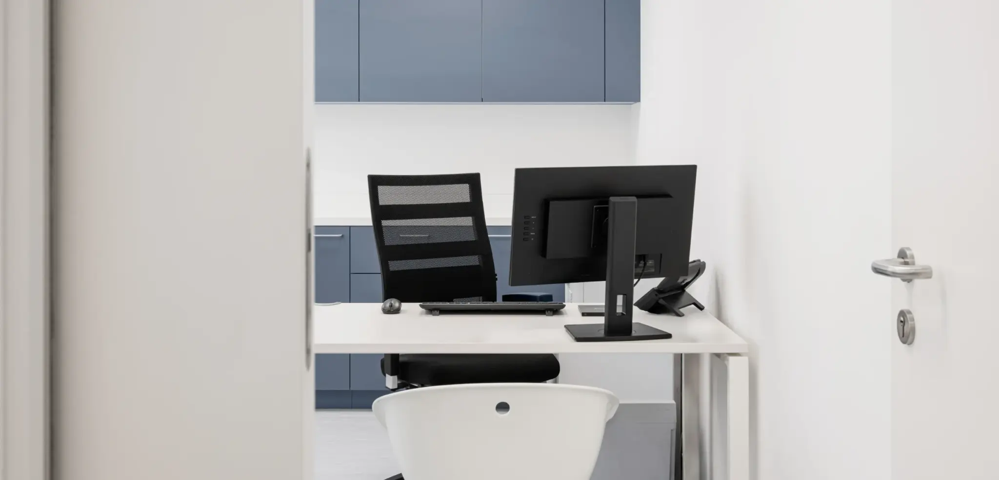 Minimalistisch eingerichtetes Arztbüro mit Schreibtisch, schwarzem Drehstuhl und Monitor, Blick durch geöffnete Tür.